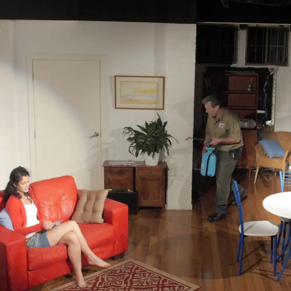A staged indoor scene: woman on red couch, man with spray canister, white walls, wooden floor.