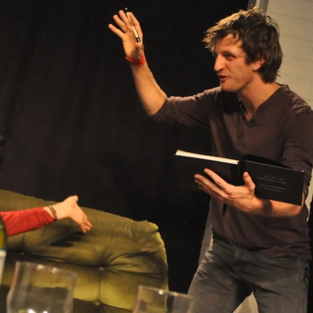 A candid photo of a man energetically gesturing with a book, next to someone reclining on a couch.