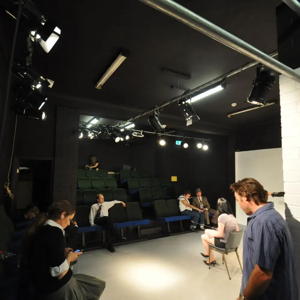 A dimly lit theater with scattered seated people, focusing on two people chatting on stage.
