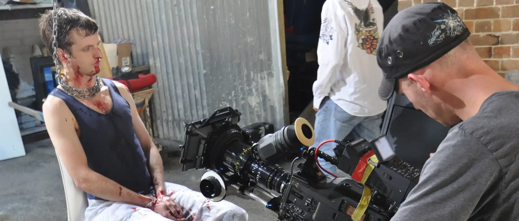 A film set showing a man with fake blood sitting, surrounded by crew and camera equipment.