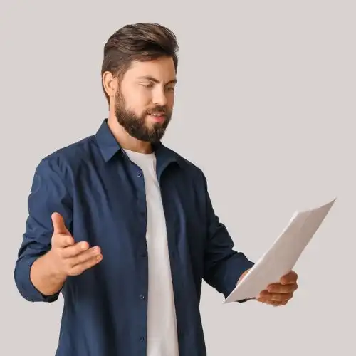 A photo of a man with a beard in a blue shirt holding a sheet of paper, gesturing with his other hand.