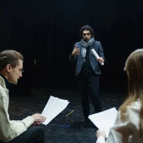 A photo of three people rehearsing on a dimly lit stage, holding papers.