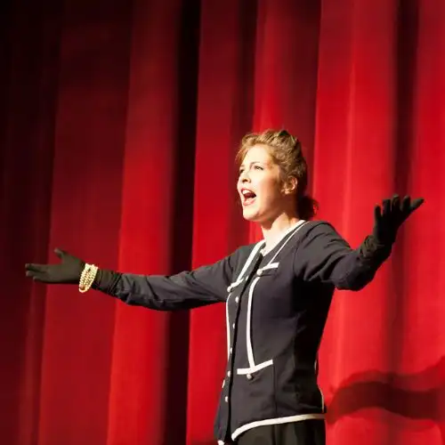 A woman on stage with a red curtain, wearing a dark outfit, arms outstretched, singing passionately.