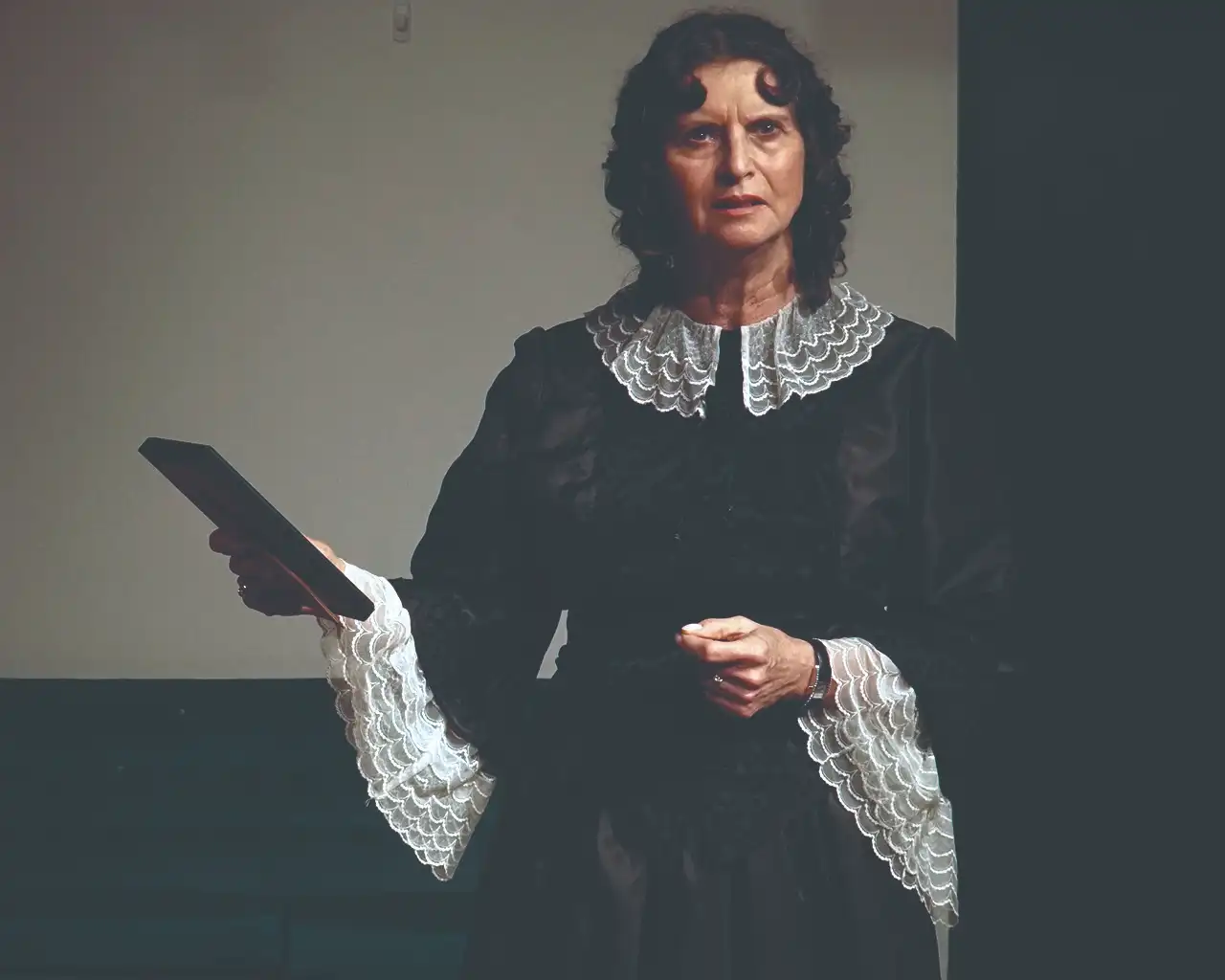 A woman in dark vintage clothing holding a book, with a serious expression.