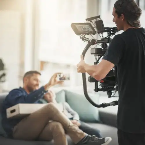 A photo of a filmmaker recording a man sitting on a sofa with a camera rig.