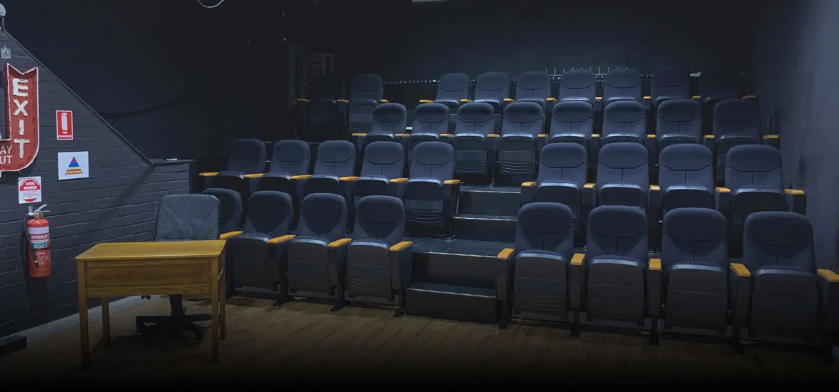 A small empty theater with tiered rows of black seats facing a wooden desk on a stage.