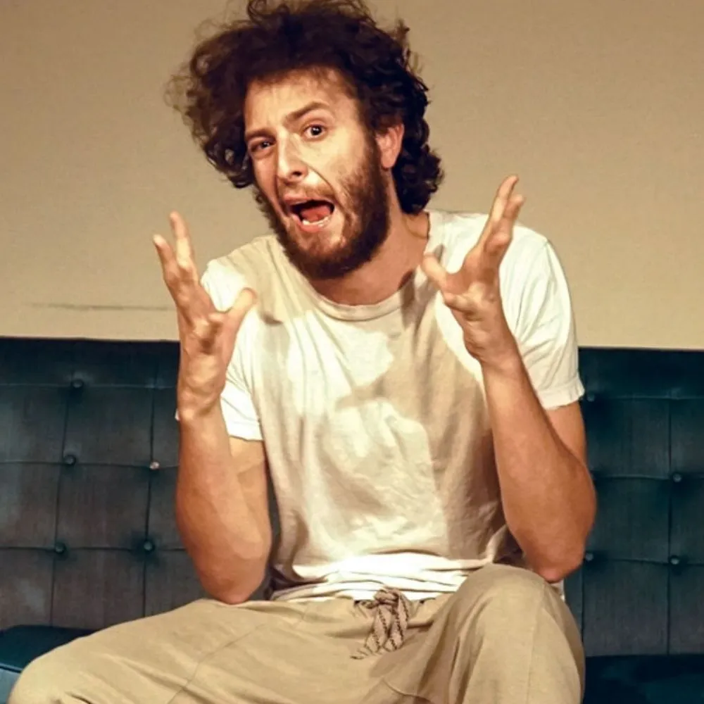 Man with curly hair and beard, wearing a white shirt, making an expressive gesture while seated on a dark couch.