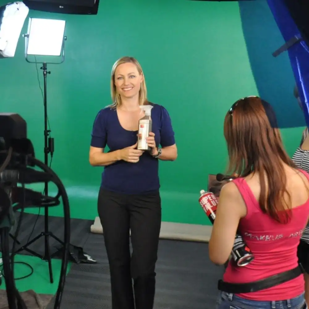 A woman in a blue top holds a product, with a green screen and lights in the background.