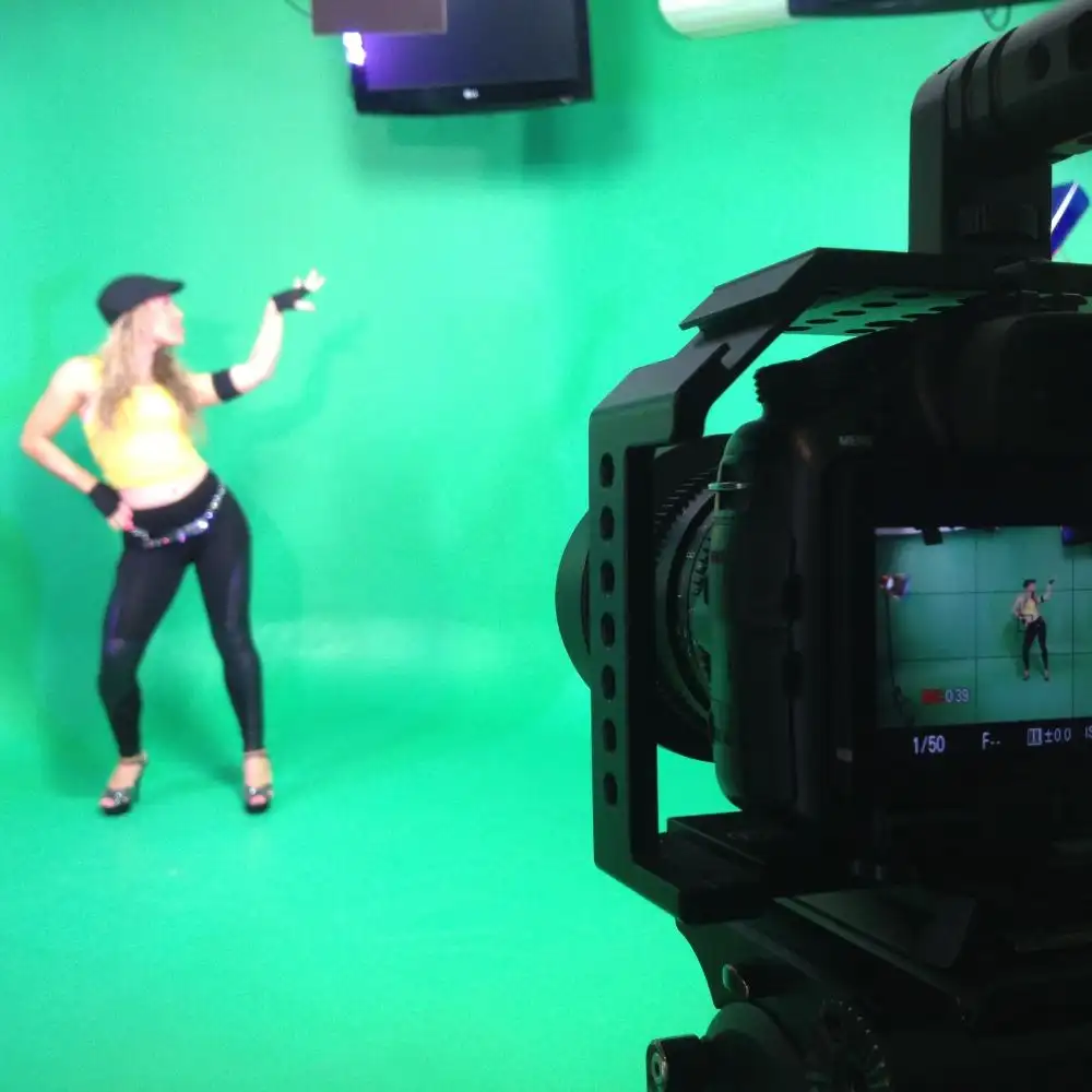 A woman poses in front of a green screen; a camera captures her.