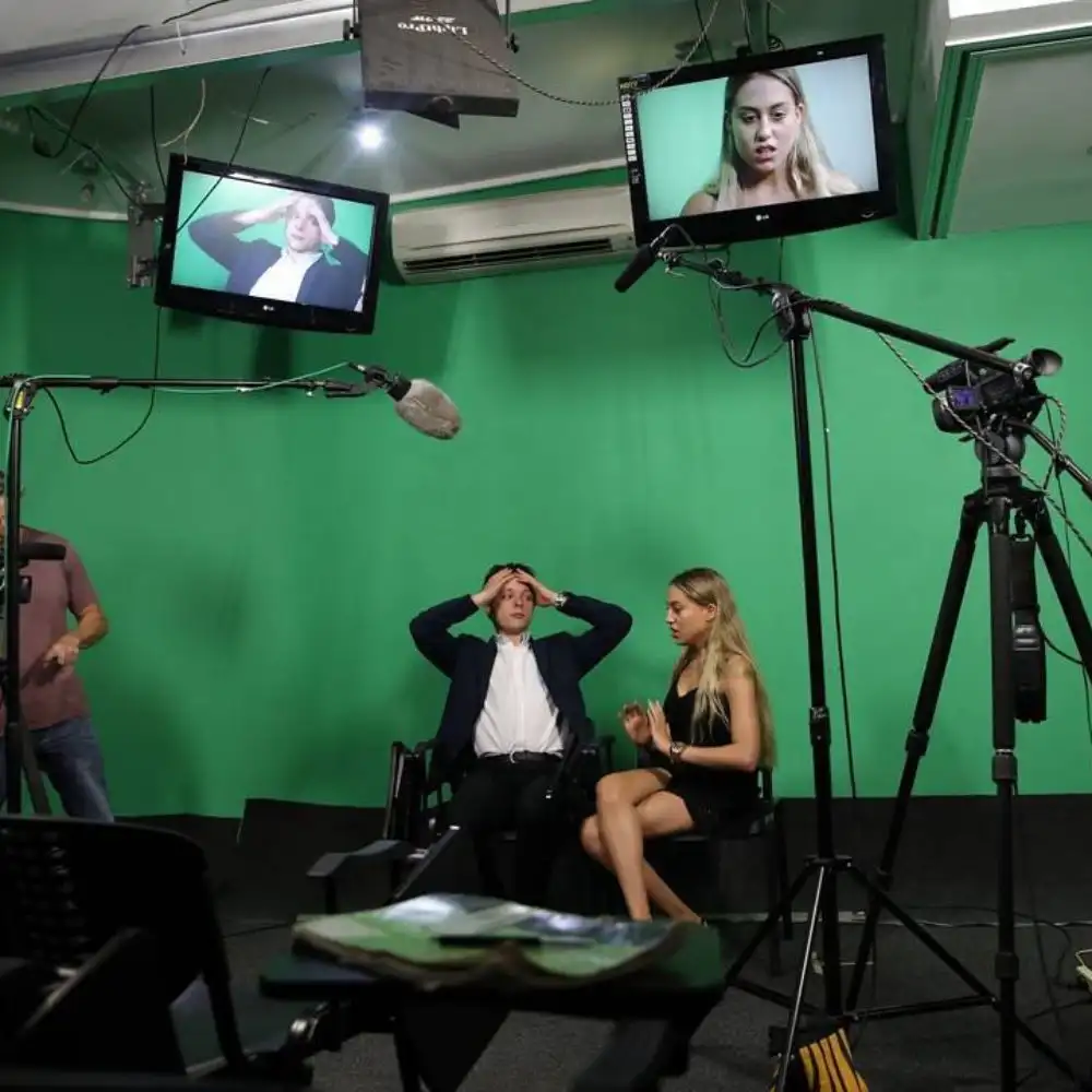 A photo of a TV studio with two people sitting, surrounded by cameras and green screens.