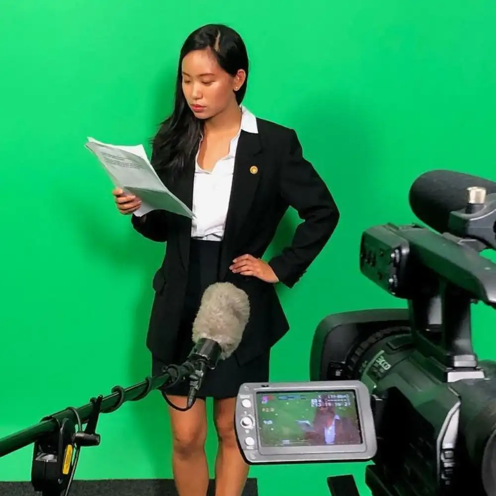 A woman in formal wear reads papers, standing in front of a green screen with a camera and microphone nearby.