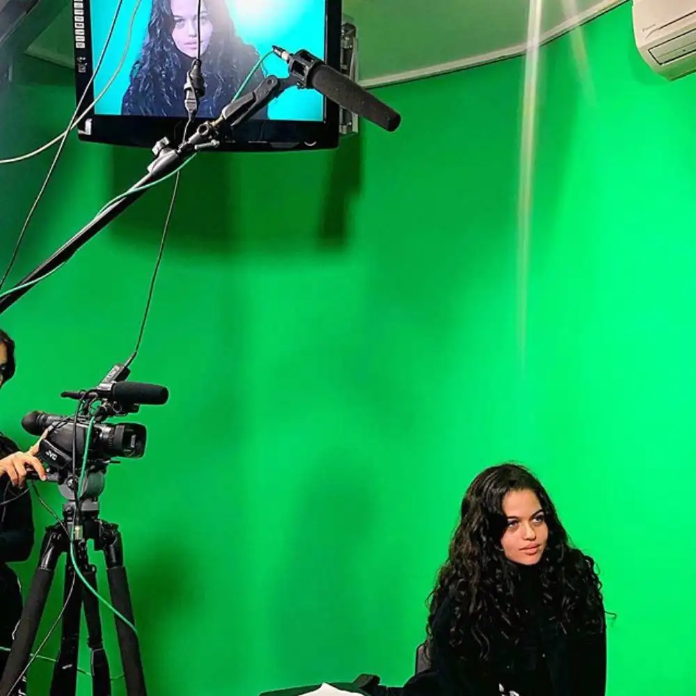 Photo of a woman with long curly hair in a green screen studio with a camera and monitor displaying her image.