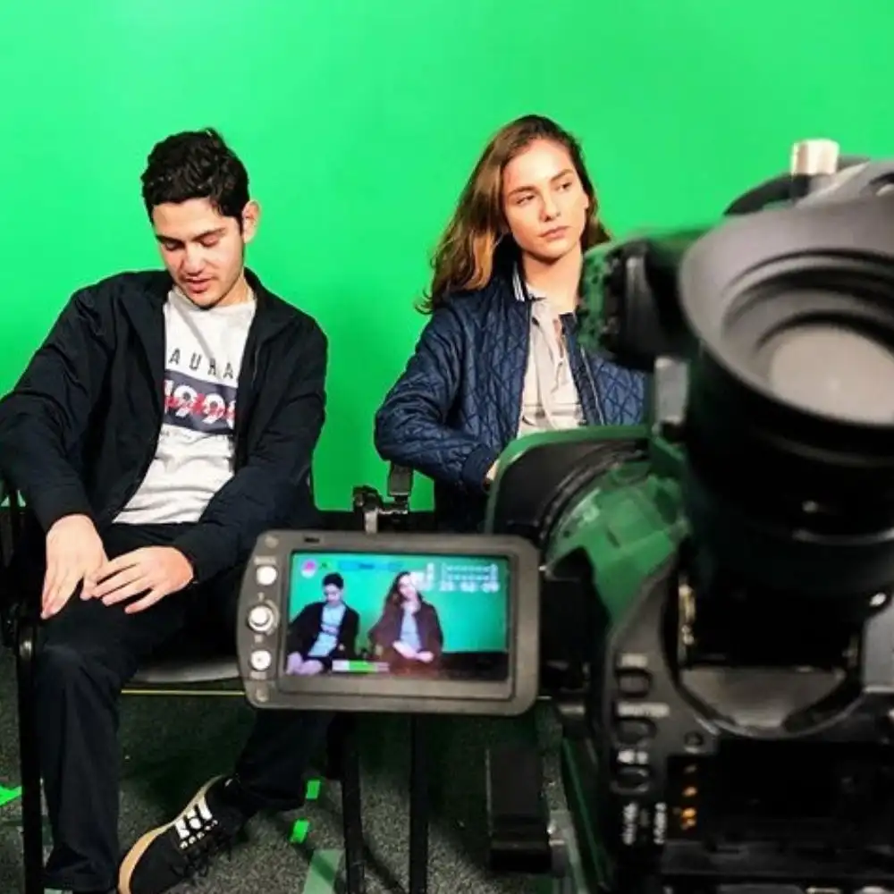 A photo of two people seated against a green screen with a camera in the foreground.