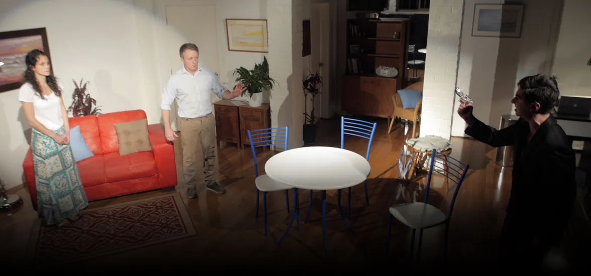 A staged dramatic scene with three people around a dining table.