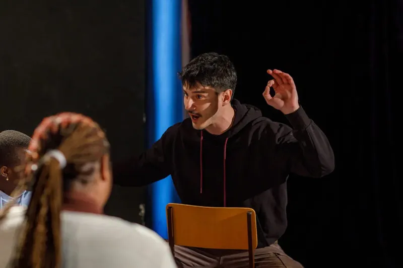 A photo of a man gesturing animatedly, seated on a chair, with two people in the foreground.