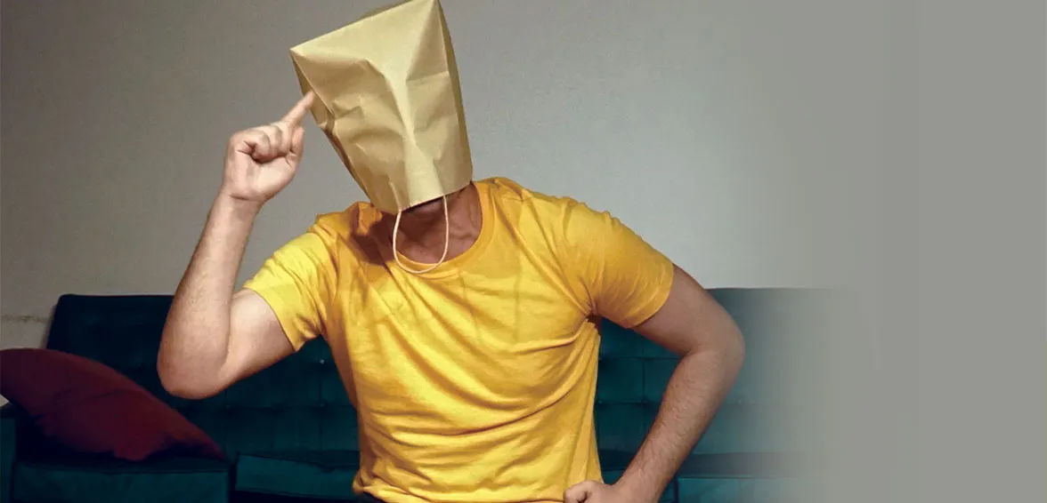 A photo of a person wearing a yellow shirt with a paper bag over their head, raising a finger.
