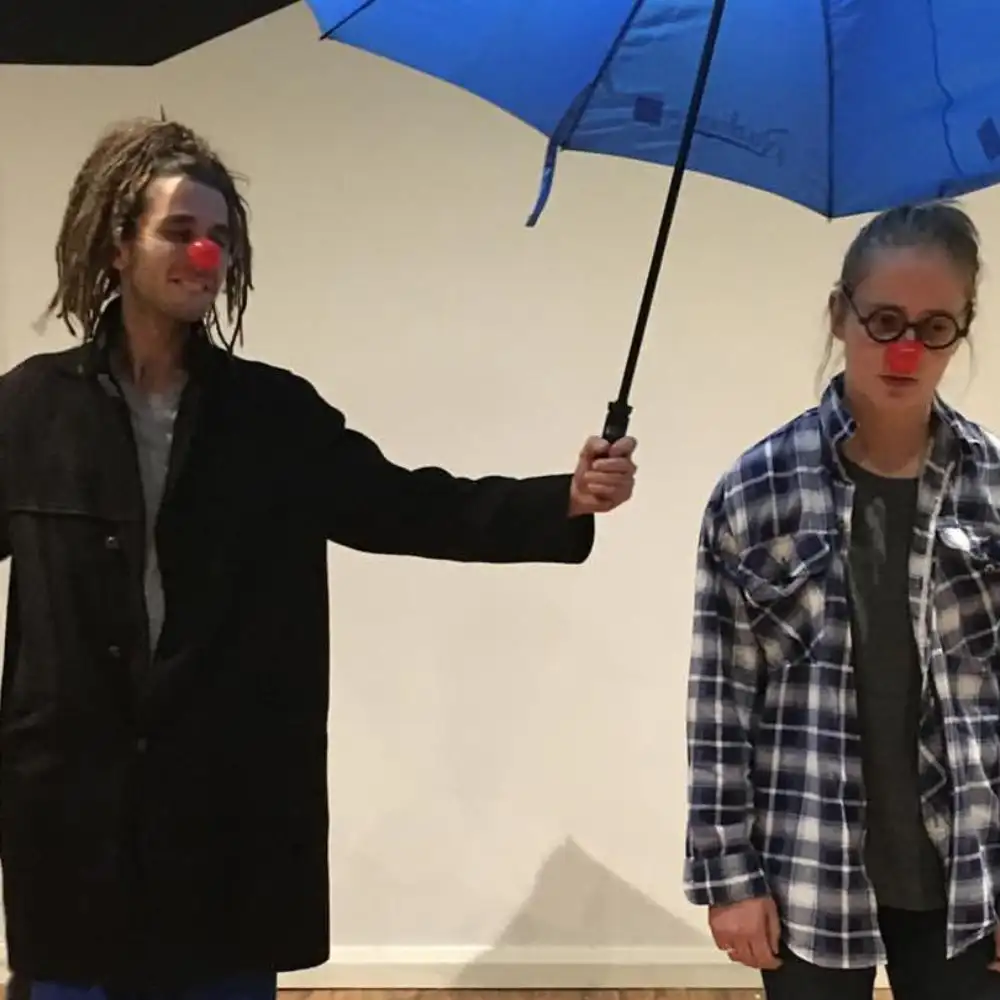 Photo: Two people with red clown noses. One holds a blue umbrella over both. Subdued expression. Casual clothing.