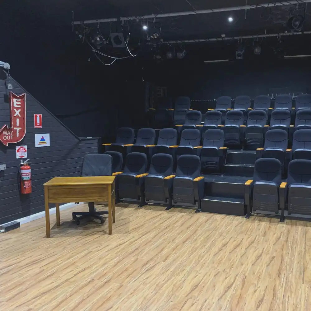 Photo of a small theater with tiered seating, a wooden floor, and a desk with a chair onstage.