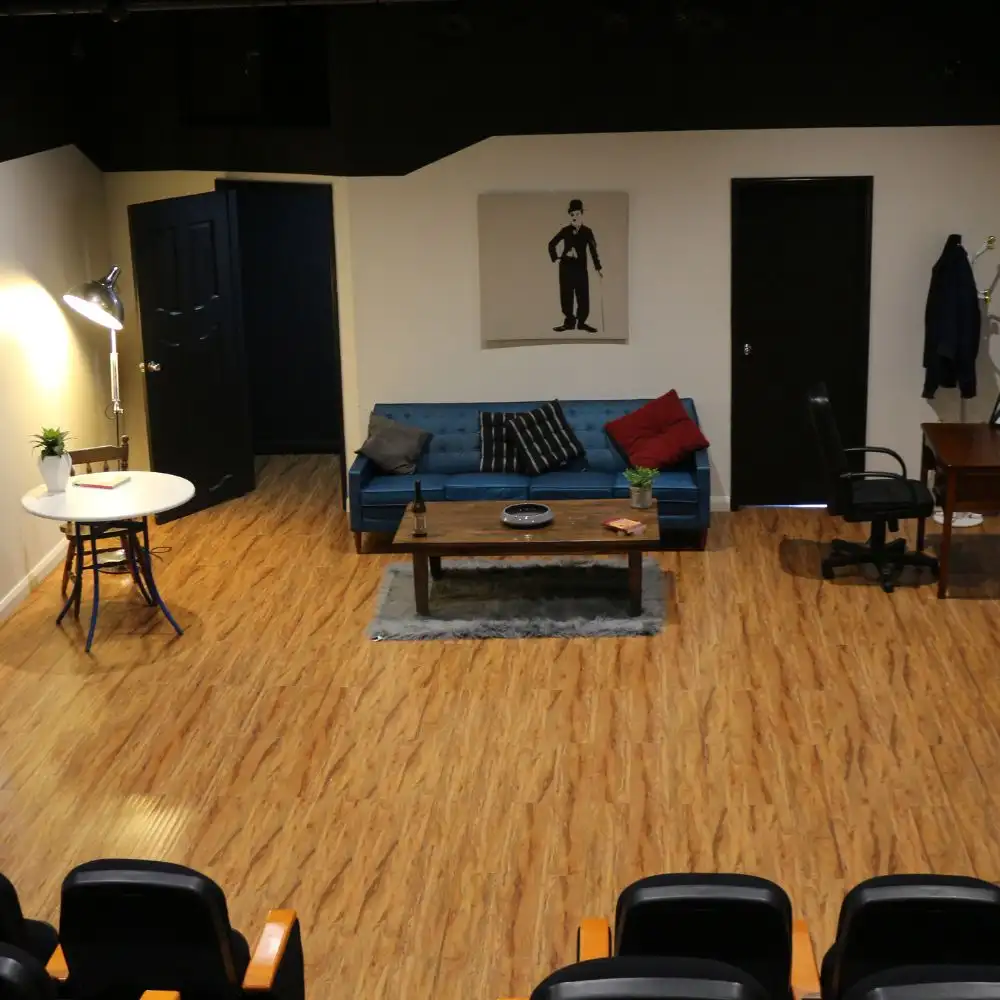 A theater stage set with a living room scene: sofa, table, chairs, doors, and wall art.