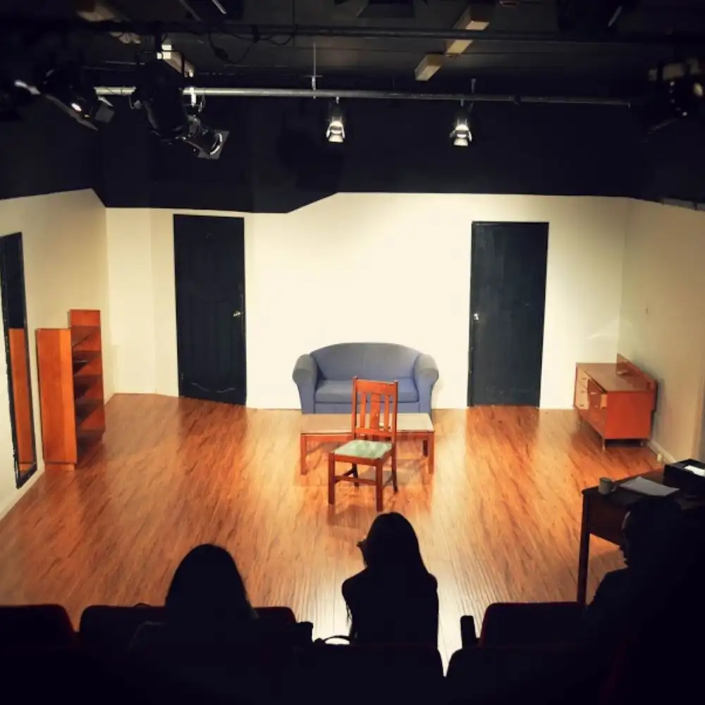 Photo of a small, empty theater stage with two doors, a blue sofa, a wooden chair, and a few shelves.