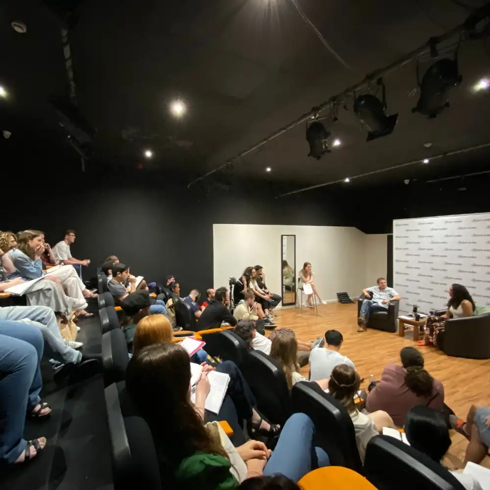 A photo of a group seated in a small theater, watching a panel discussion on a stage with three speakers.
