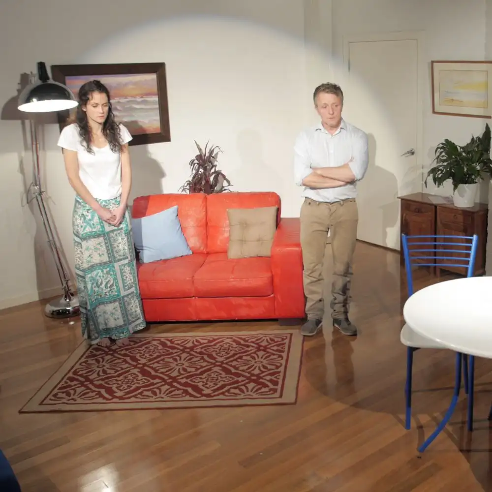 Photo: A woman and a man stand in a living room with a red sofa, rug, plants, and a round table.