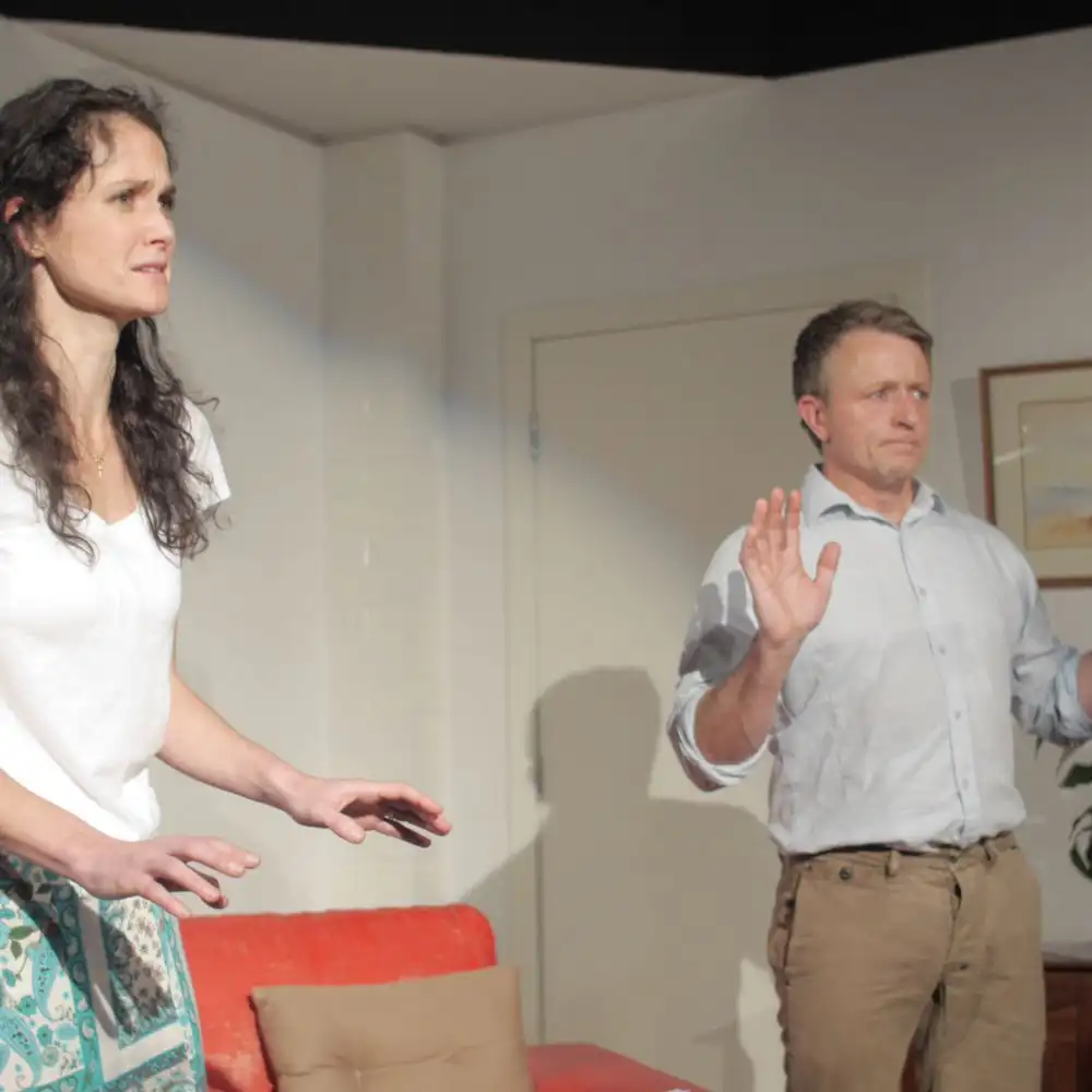 A man and a woman in a room with a red sofa, both have their hands raised.