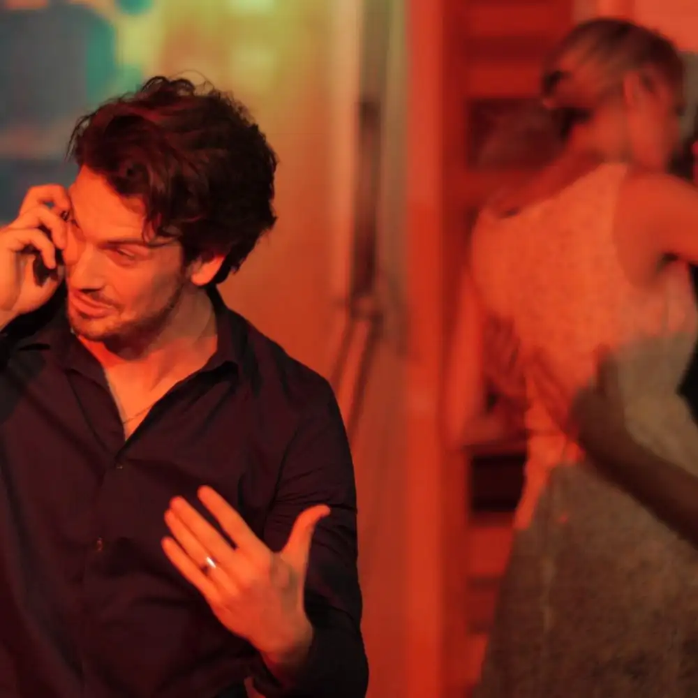 A photo shows a man talking on the phone in a dimly lit room with a woman blurred in the background.