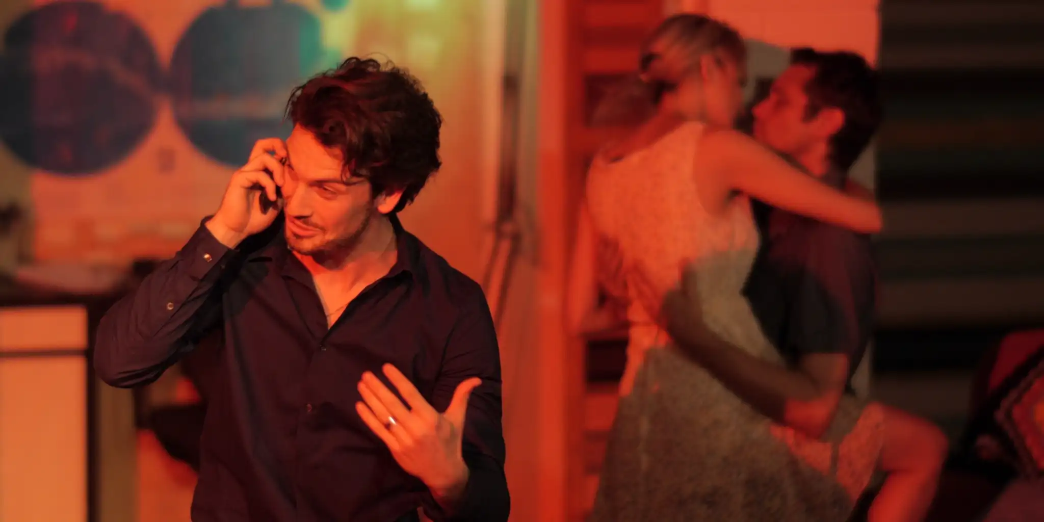 A man on the phone looks upset, while a couple embraces in the background. Warm, dim lighting. Color photo.