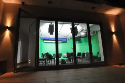 Photo of a room with a green screen, chairs, and tables, viewed through glass doors.
