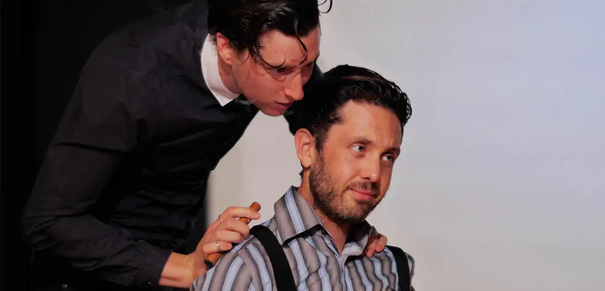 Two men, one standing and leaning in, the other sitting, looking apprehensive. Indoors, casual clothing.