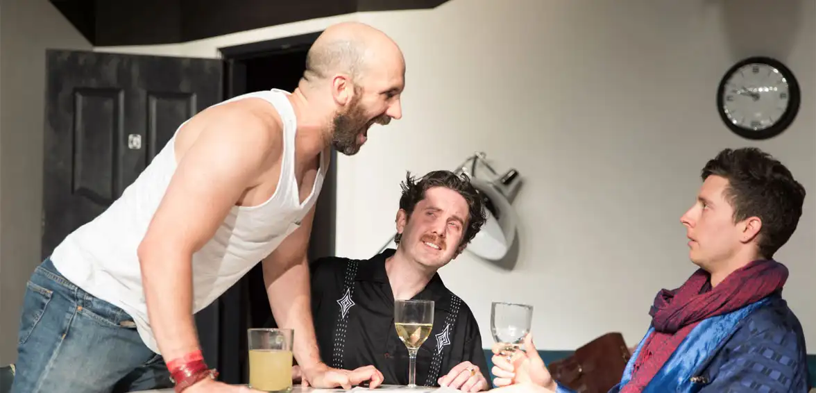 A photo of three men at a table with drinks. One man is standing and yelling, while two others sit and listen.