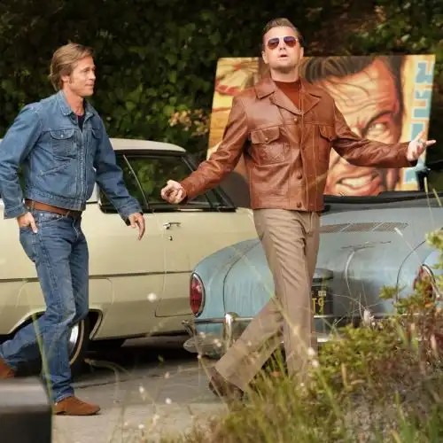 A scene from a movie with two men walking near vintage cars. One wears denim, the other a brown leather jacket and sunglasses.