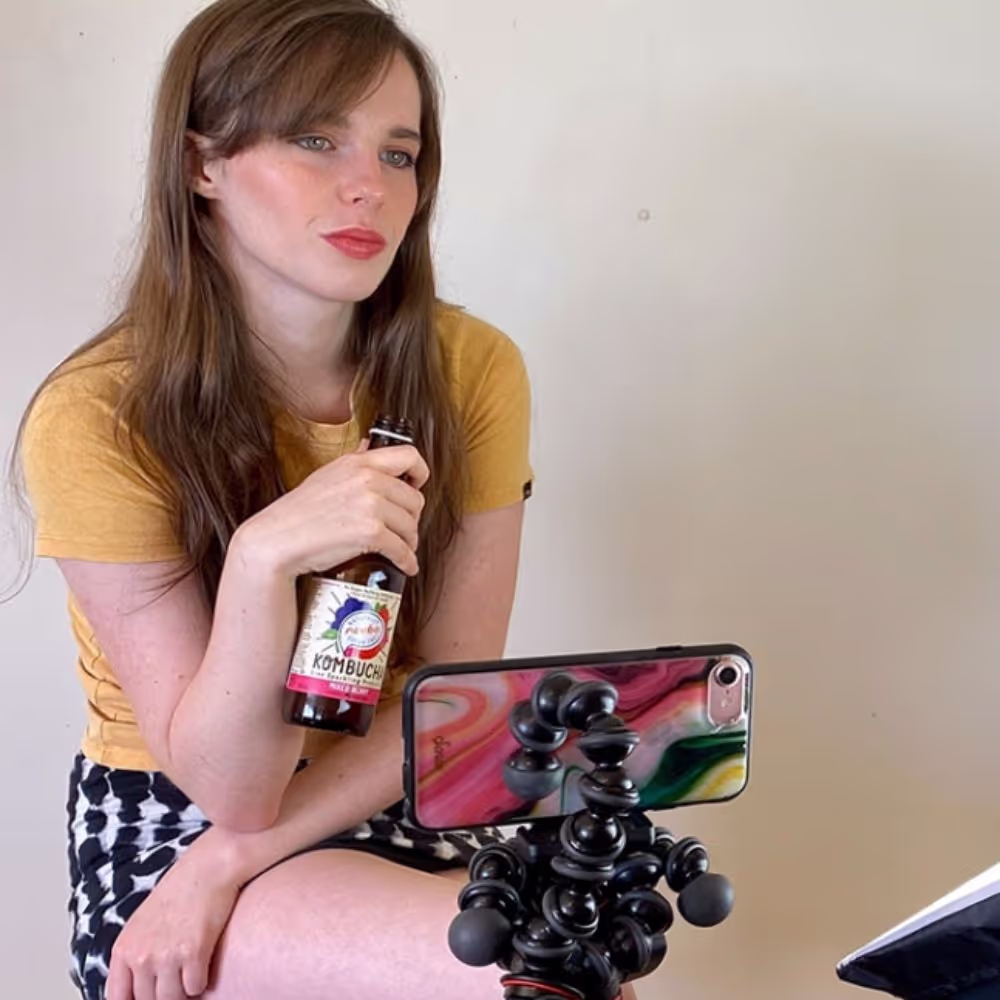 Young woman with long brown hair, wearing a yellow shirt and patterned skirt, holding a bottle of kombucha while sitting in front of a phone mounted on a flexible tripod.