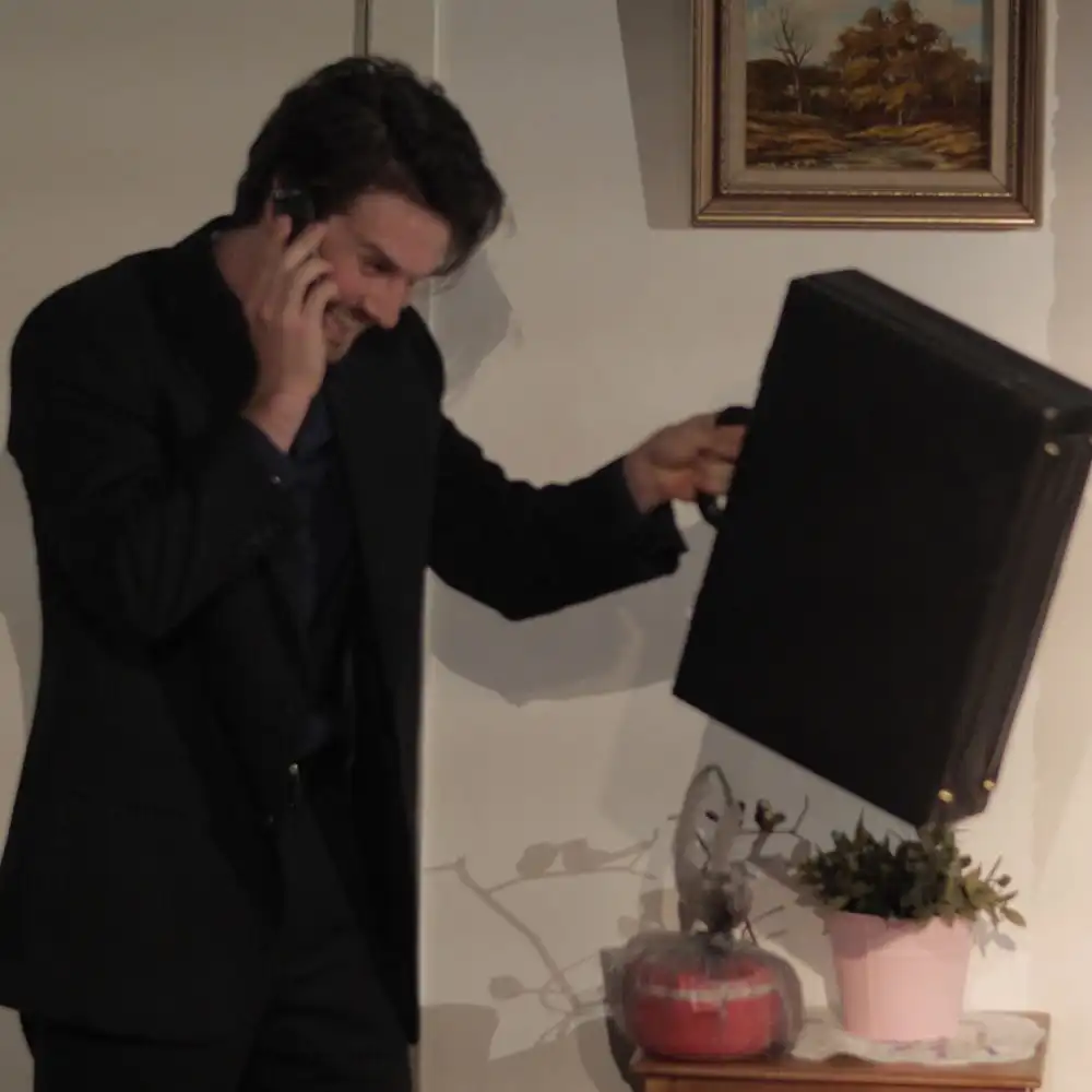 Man in a black suit talking on the phone and holding a black briefcase in a room with a plant and a painting on the wall.