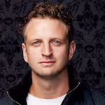 Portrait of a young man with short curly hair and blue eyes wearing a black jacket over a white shirt against a dark patterned background.
