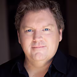Headshot of a man with light skin, blue eyes, and short brown hair wearing a dark shirt against a dark background.